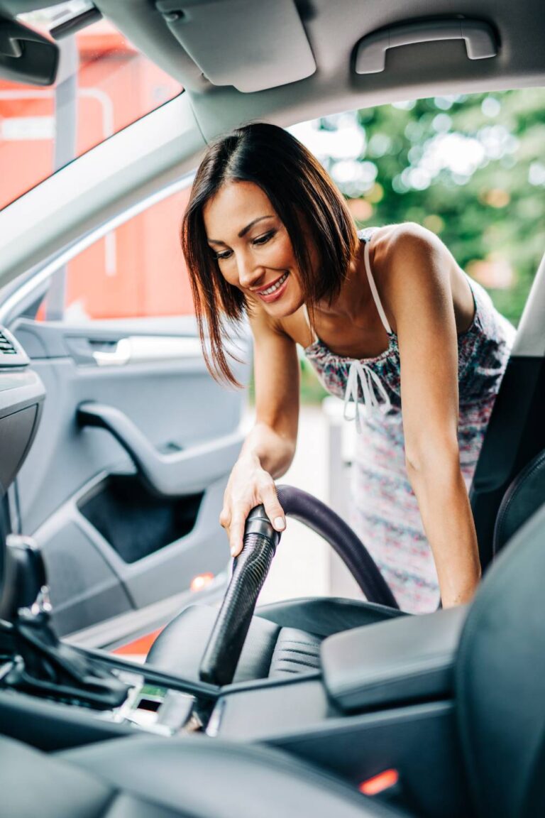 A smiling woman in a long dress leans into her car on the passenger side to vacuum between the seat and console.