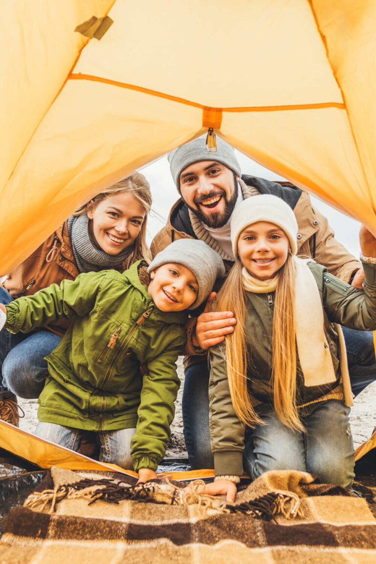 Two adults and two children wearing winter jackets and looking into the entrance of a yellow tent, smiling.