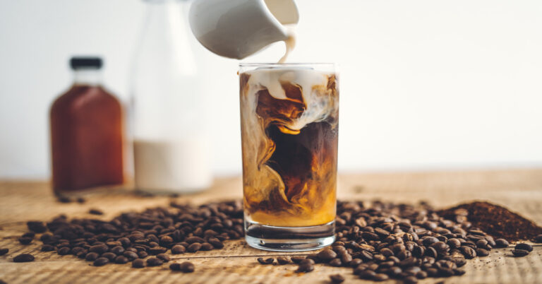 A glass with cold brew coffee and whole coffee beans scattered around the glass. Someone is pouring cream into the glass.