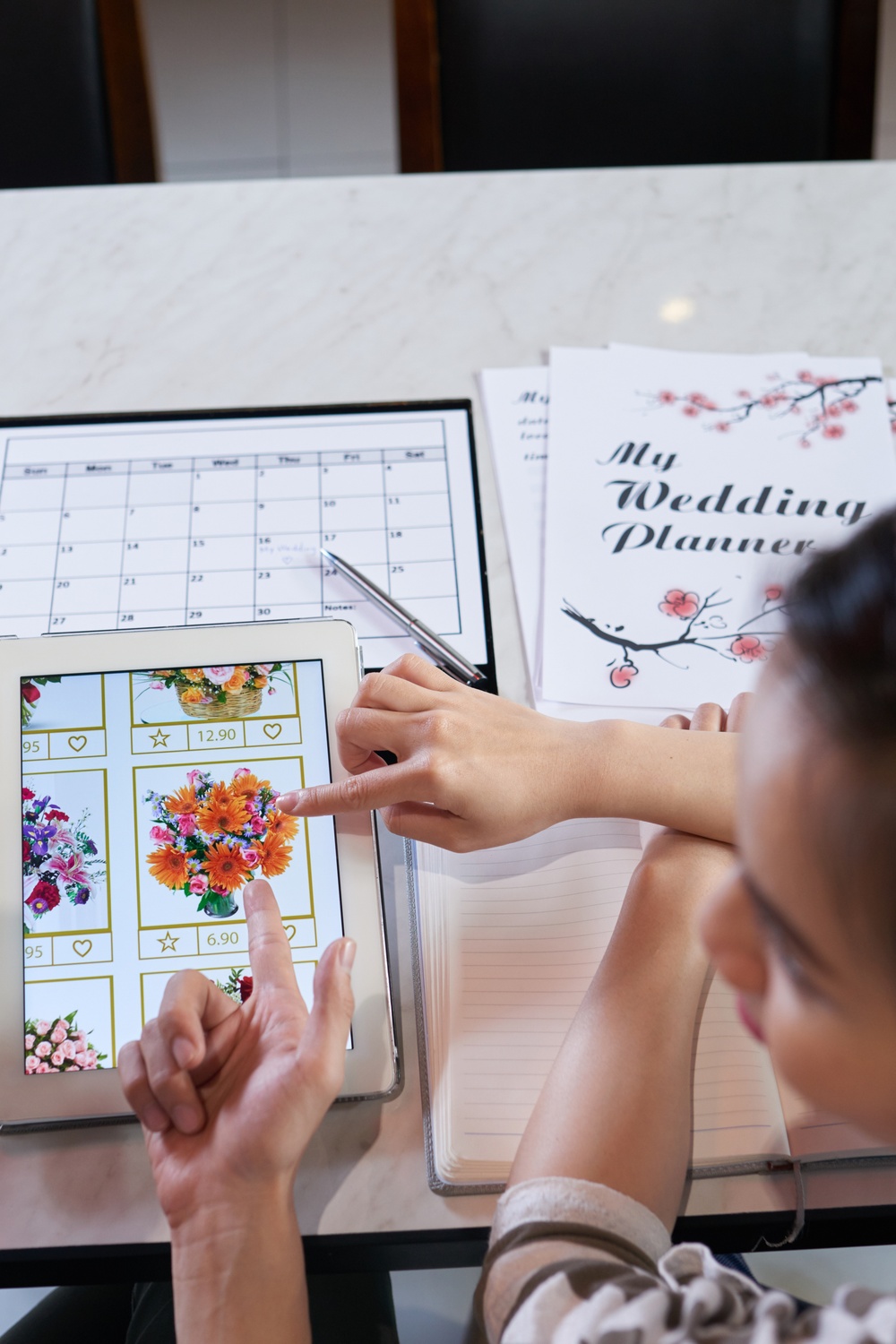 An overhead view of a man and woman looking at pictures of flowers on a tablet next to a calendar and planning notebook.