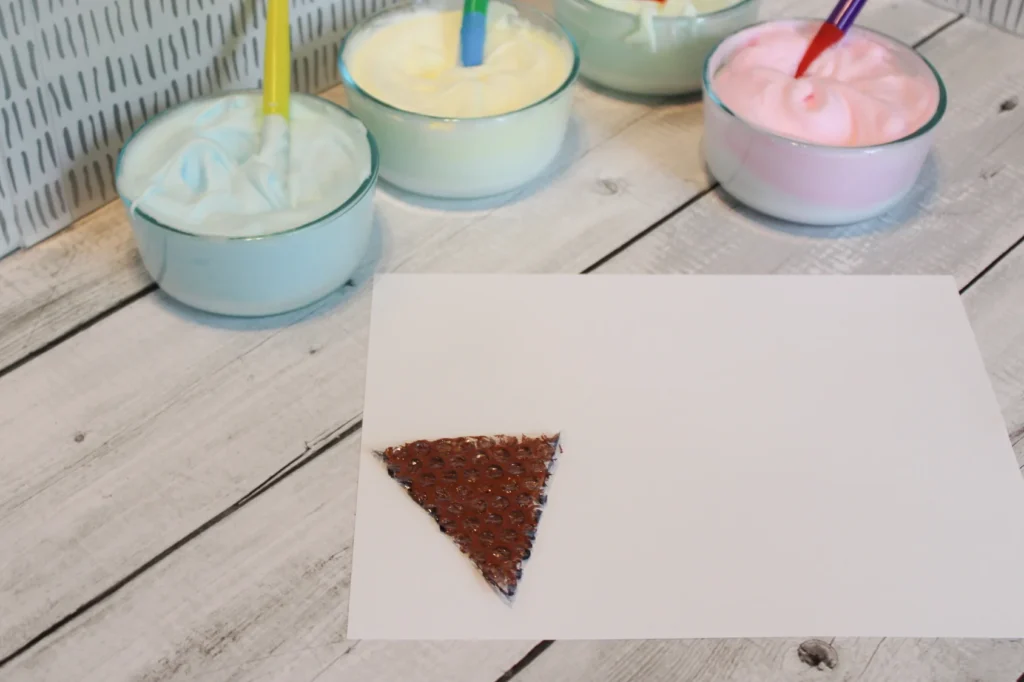 Brown stamped triangle cone on paper with puffy paint in bowls and brushes ready to decorate.