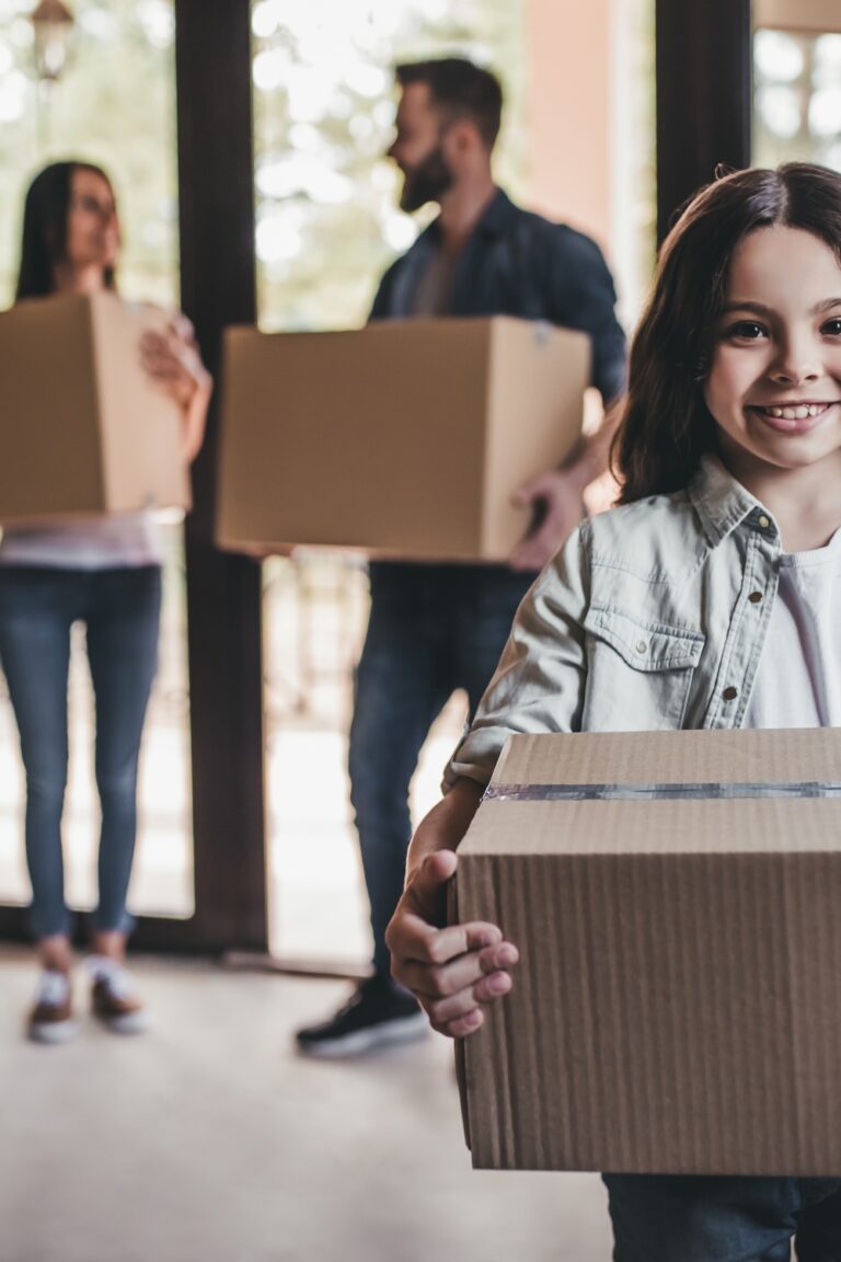 A young girl holds a moving box and smiles toward the camera. A man and woman stand behind her, blurred.