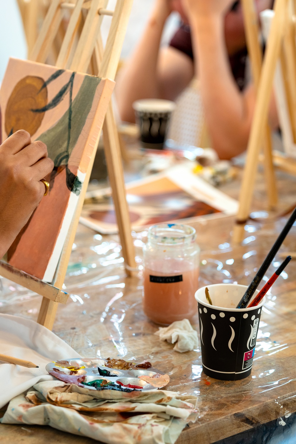 A woman’s hands painting on a small canvas on an easel, with other people painting together in the background.