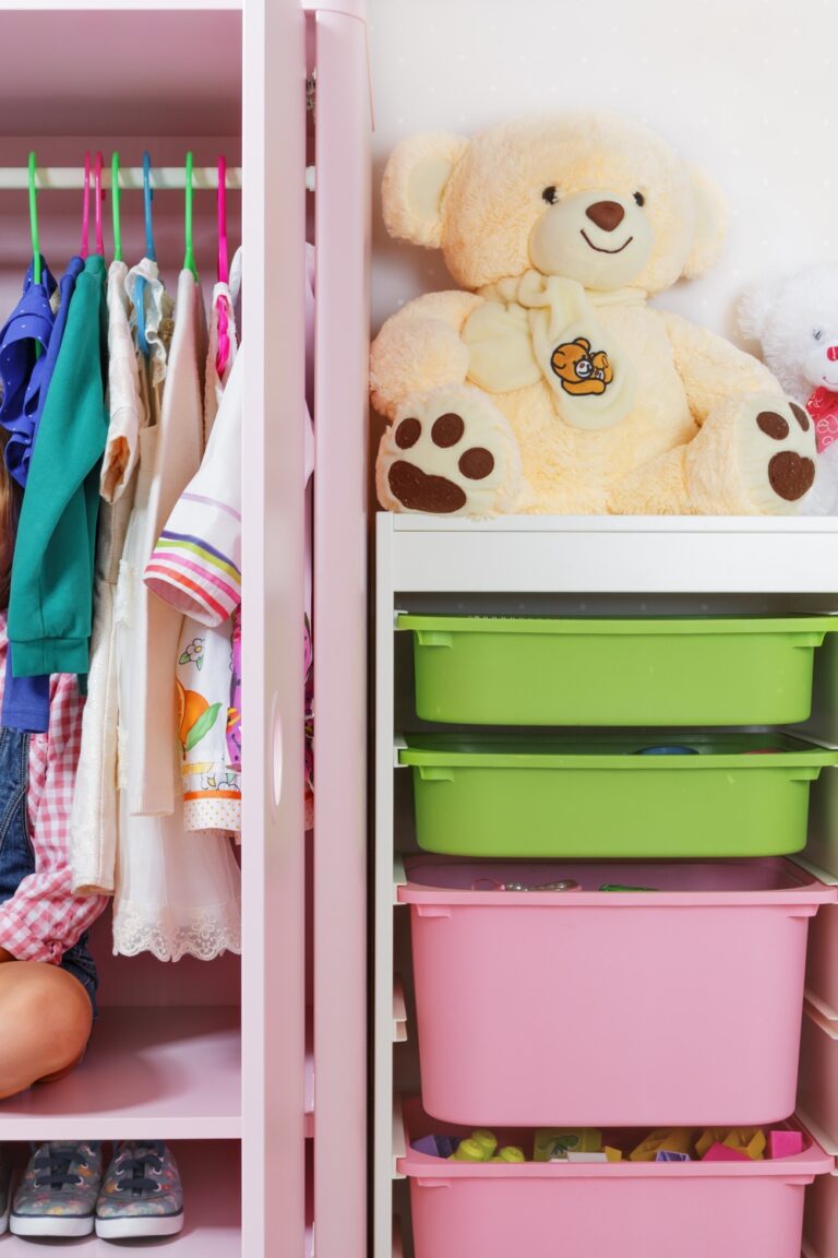 A small girl sits on the upper shelf of her open pink closet. She's in between clothes hanging from colorful hangers.