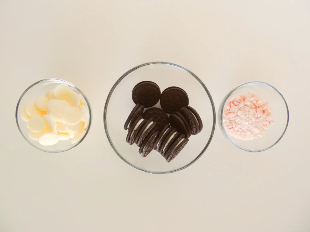 Three glass bowls holding white chocolate melting wafers, Oreo cookies, and crushed peppermint candy on a white background.
