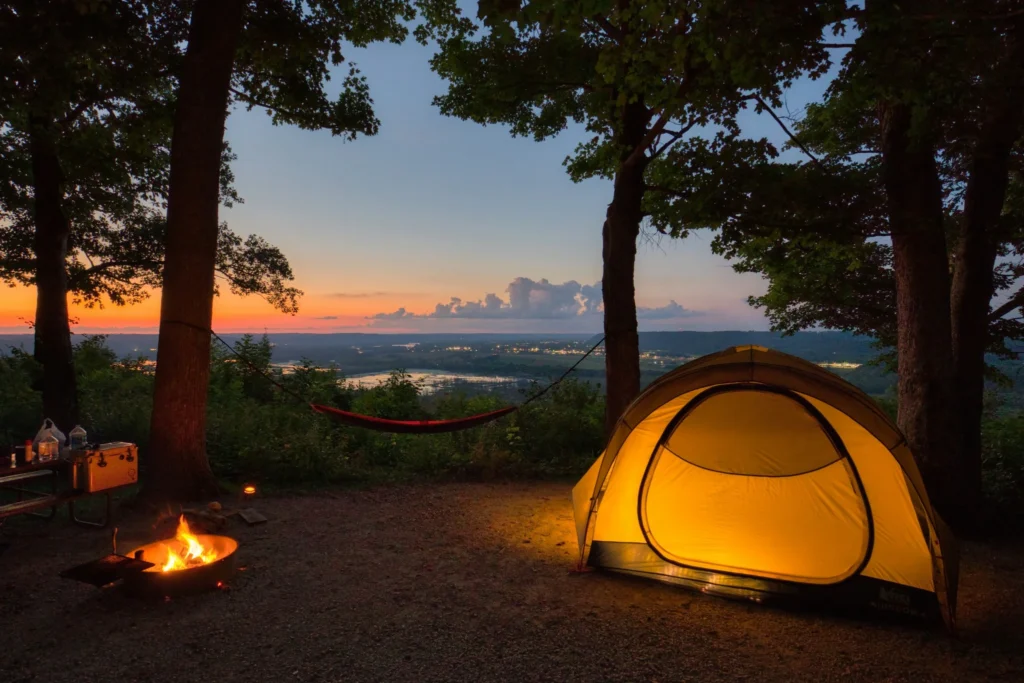 A glowing camping tent sits beside a campfire at sunset overlooking a scenic valley.