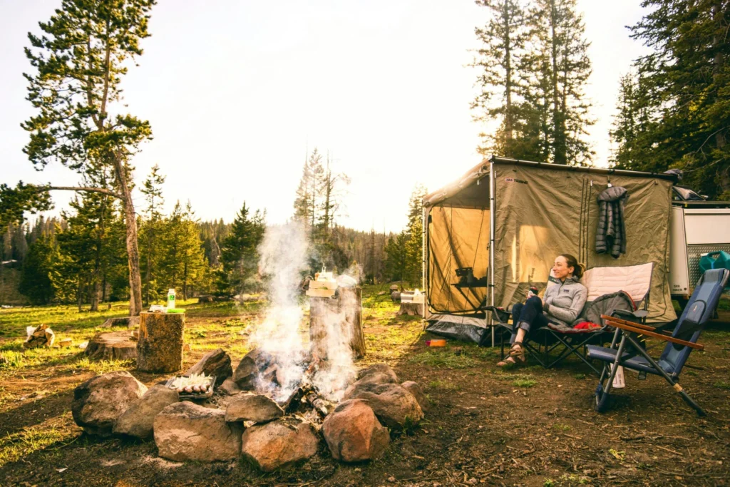 Campsite with a campfire burning in a stone fire ring beside a tent and camping chairs in a forest.