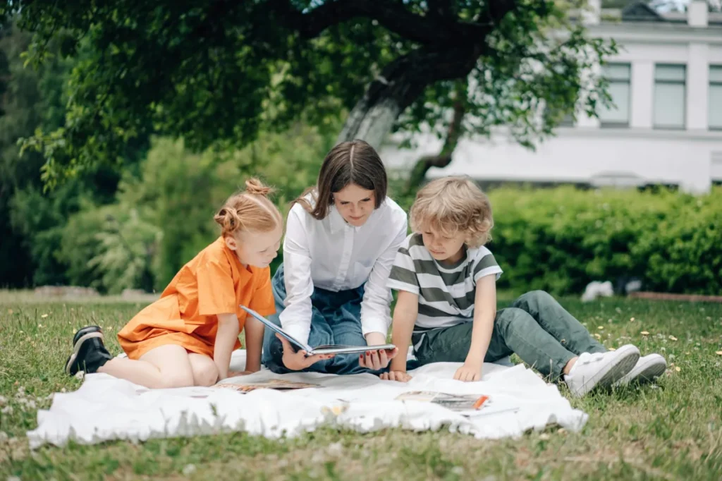 Adult reading with two children on a blanket outside in a grassy park during summer.