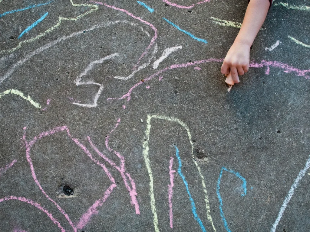 Child drawing colorful designs with sidewalk chalk on pavement outdoors.