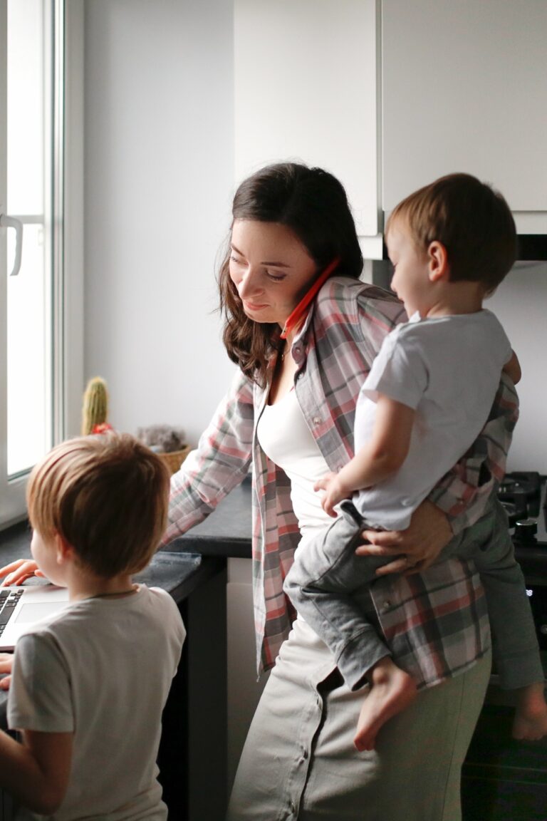 A woman holding a child while using a laptop as another child stands nearby at a kitchen counter beside a window.