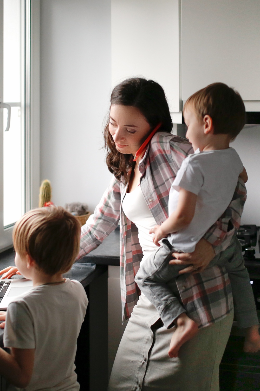A woman holding a child while using a laptop as another child stands nearby at a kitchen counter beside a window.