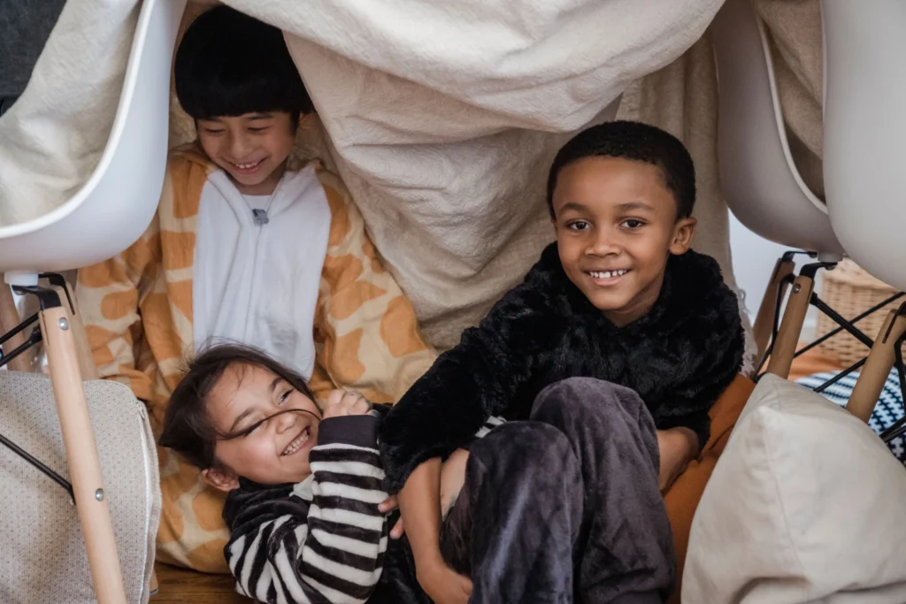 Children sitting inside a cozy blanket fort during a rainy day indoors.