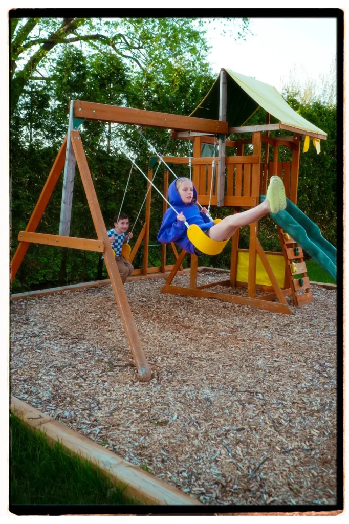 Two children playing on a wooden backyard swing set with a slide and climbing wall