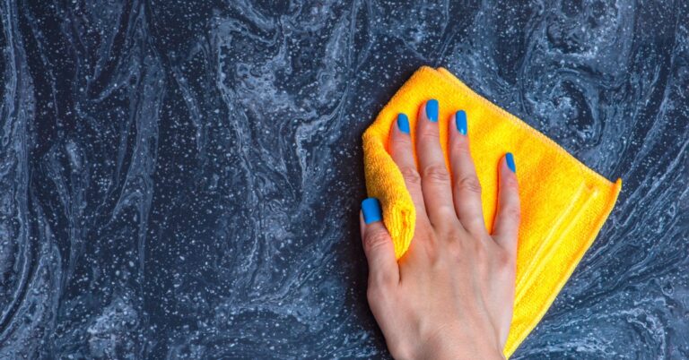 A close-up of a hand with blue fingernails wiping a black and blue stone countertop with a yellow rag.