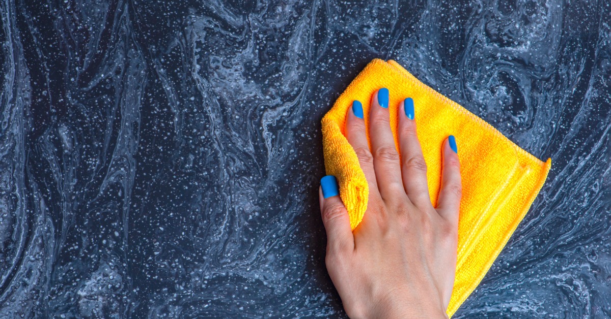 A close-up of a hand with blue fingernails wiping a black and blue stone countertop with a yellow rag.