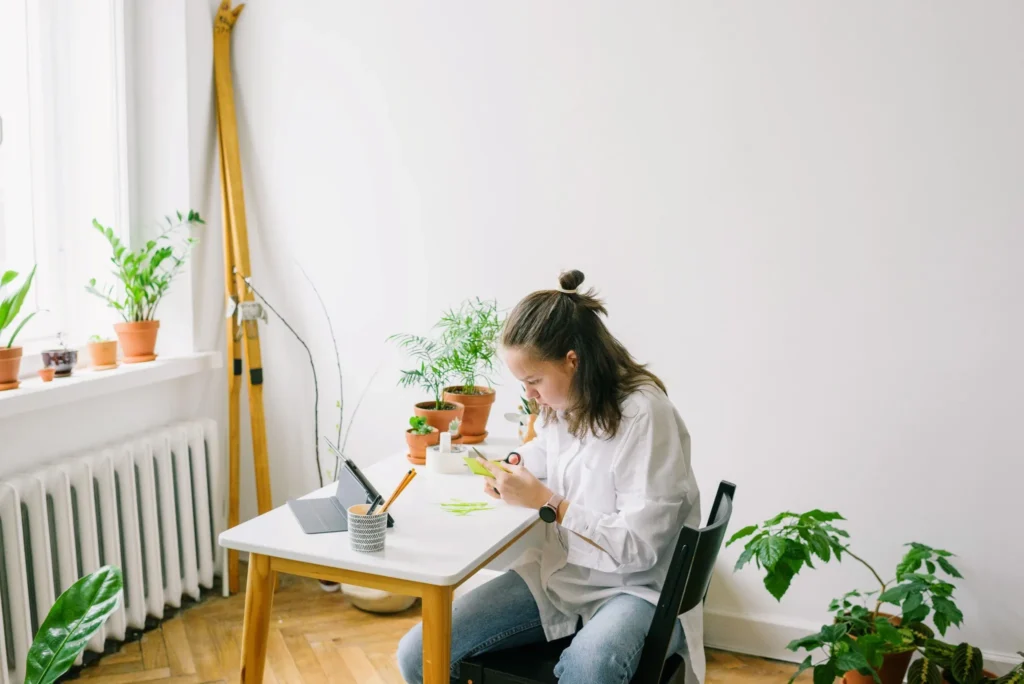 Woman working on a paper craft at a table beside a large window with natural light