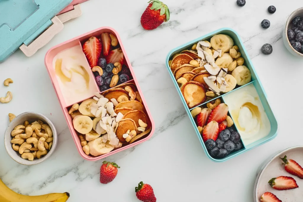 Two finished breakfast lunchboxes with mini pancakes, yogurt, fresh fruit, cashews, and coconut