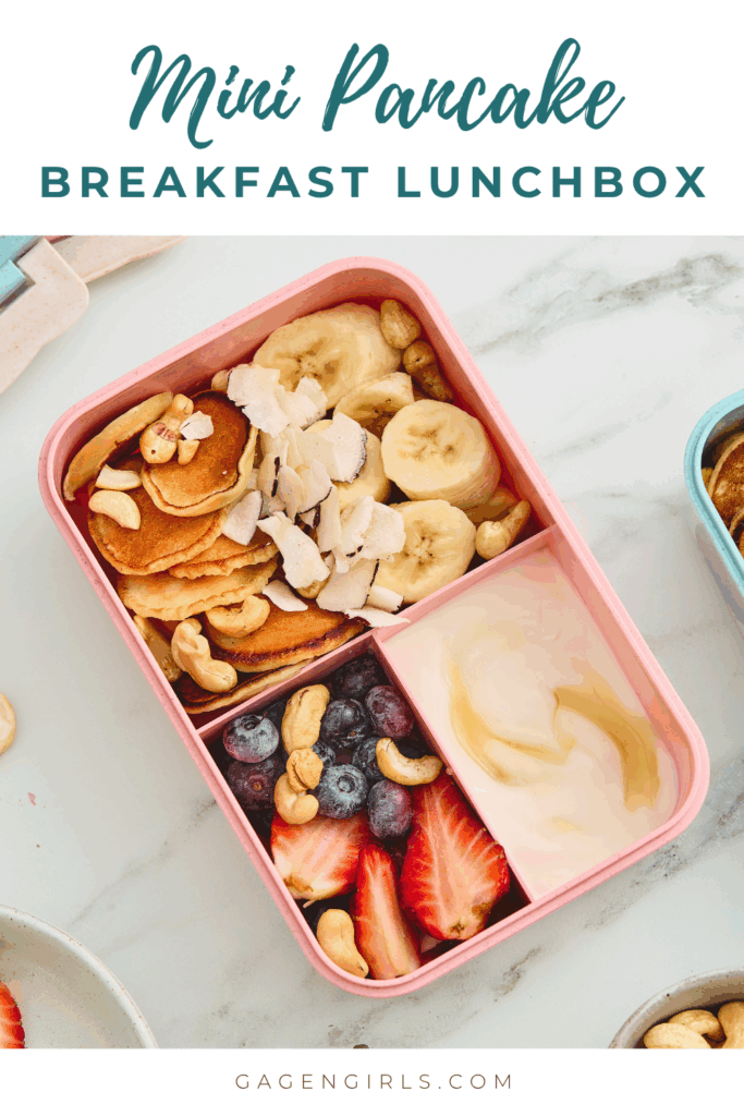 Pink breakfast lunchbox filled with mini pancakes, banana, strawberries, blueberries, yogurt, cashews, and coconut.