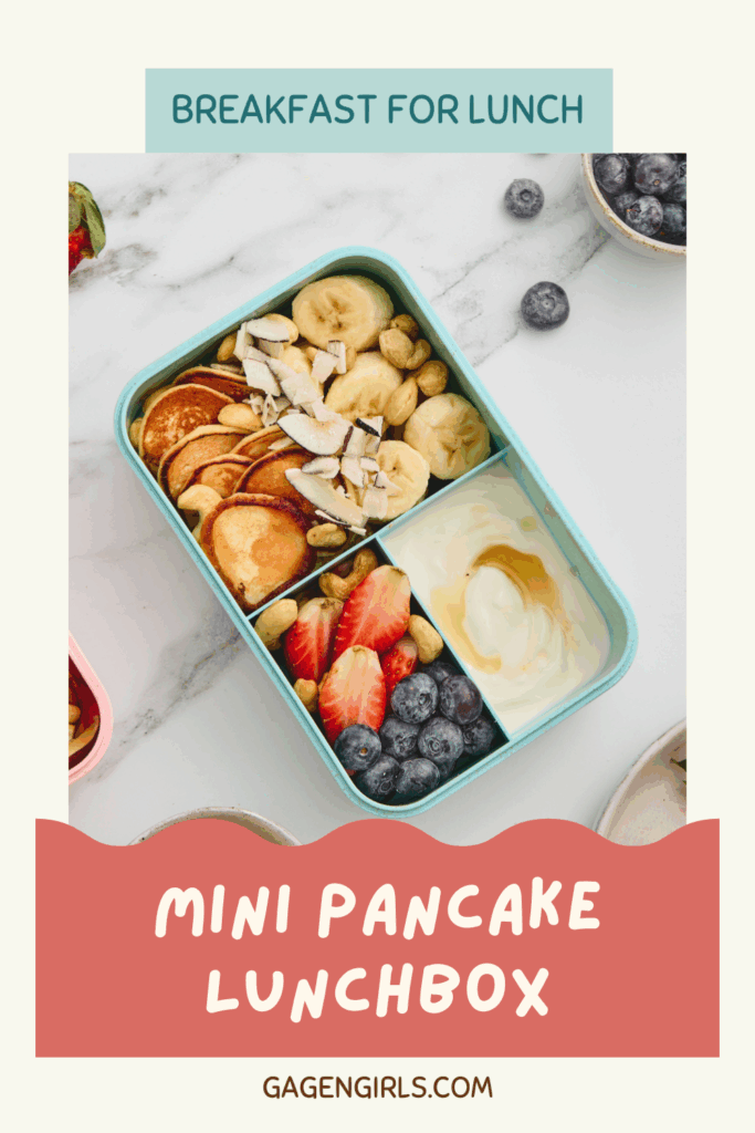 Teal lunchbox containing mini pancakes, banana slices, strawberries, blueberries, yogurt, cashews, and coconut.