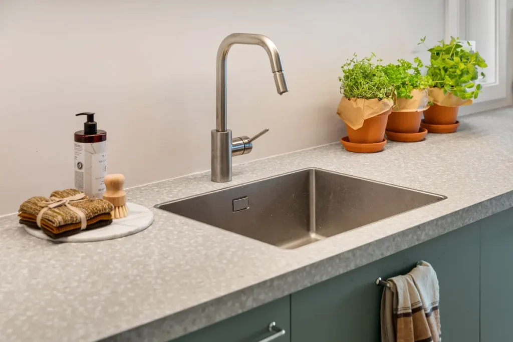 Kitchen sink set into a light-colored countertop with soap, herbs, and cleaning cloths nearby.