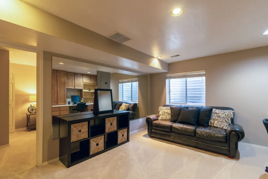 Multifunctional basement living area with a sofa, home office nook, storage shelves, and windows providing natural light.